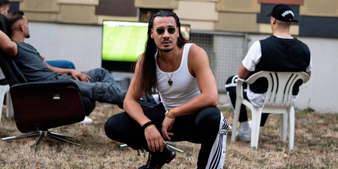 Ein Mann mit längeren dunklen Haaren, weißem Tanktop und Sonnenbrille hockt im Vordergrund. Dahinter schauen ein paar Männer in Plastikstühlen Fußball auf einem Fernseher.